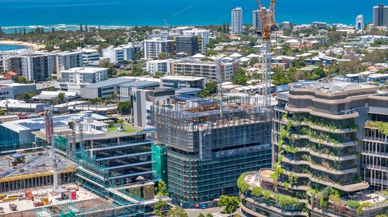 50 First Avenue under construction as seen from a distance. Nearby smaller apartment buildings and houses are visible in the distance, after which the wide blue ocean extends out to the horizon.