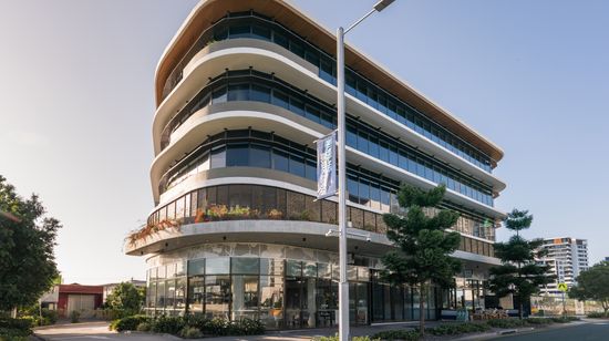 A1 Maroochydore seen from street level, with warm sunlight reflecting off its wraparound glass windows. The taller ground floor has several restaurants and cafés seen through the glass walls.