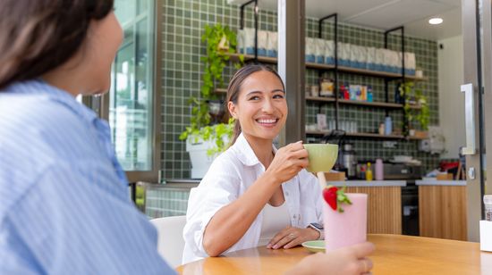 Two friends sitting at a café’s outdoor table. One person is smiling while enjoying a coffee, listening to the other who is having a drink garnished with a strawberry.