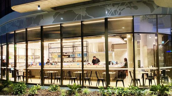 The bright interior of the restaurant Kibou in A1 Maroochydore’s ground floor, seen through its glass walls from outside at night. Several two-person tables line the walls around the open kitchen with a wraparound counter in the centre of the restaurant.