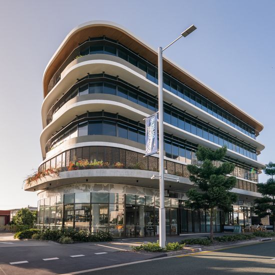 A1 Maroochydore seen from street level, with warm sunlight reflecting off its wraparound glass windows. The taller ground floor has several restaurants and cafés seen through the glass walls.