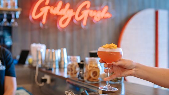 A pink cocktail garnished with a yellow flower being held at the bar at the restaurant Giddy Geisha within Foundation Place. A neon-style “Giddy Geisha” sign is seen slightly out of focus on the wall in the background in the same colour as the drink.