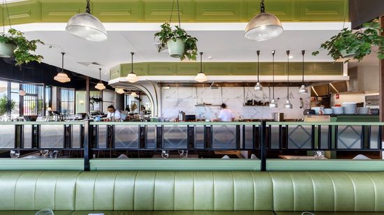 The interior of a modern restaurant with striking mint green accents. The lounge seating areas and roof accents are all a matching shade of green. In the background staff can be seen preparing in the open kitchen.