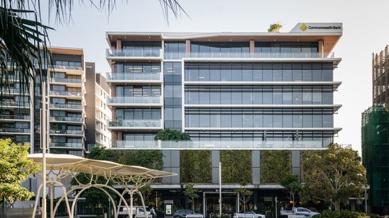 The front of Foundation Place seen from across a neighbouring grassy field. The tall ground floor has a large entrance to the lobby, surrounded by several retailers. Large green walls span the next floor, leading to several floors of modern glass façades. The Commonwealth Bank’s logo is featured on the top right corner of the building.