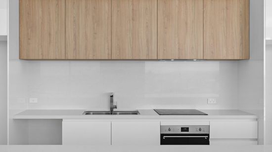 A modern kitchen of an apartment at Market Lane, with white benchtops and cabinets inset with stainless steel appliances. A light wooden cabinet is suspended above the kitchen.