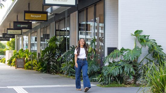A person smiling while walking past the row of retailers at the ground floor of Market Lane’s townhouses. The retailers include a salon, restaurant, real estate agent, and wellness centre.