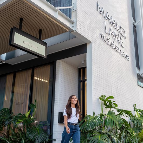 A person smiling while walking past the salon Tarli Hair in the ground floor of Market Lane’s townhouses area. A large “Market Lane Residences” sign is seen on the wall above them.