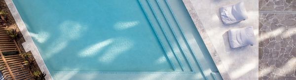The residents’ pool at The Corso seen from above. The pool is clear blue and features full-width stairs leading down into it. Various white fabric cushions are placed on the tiled deck area along the pool’s edge.