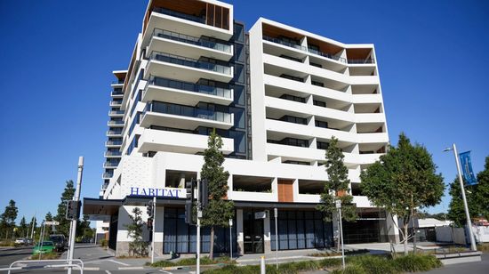 One of the apartment buildings of The Corso seen from street level. The building’s bright white façade is shining in the sunlight. The ground floor has retail spaces with a large “Habitat” sign on the awning.
