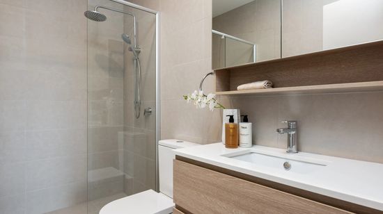 The modern bathroom of an apartment at The Corso, with sandstone coloured tiles lining the floor and walls and a white basin with wooden drawers fitted on the wall.