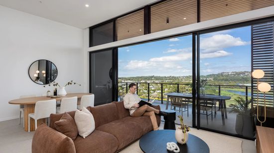 The modern living room of an apartment at The Corso. A resident is sitting and reading on a large brown couch in the centre of the room, while a designer wooden dining table with white chairs is seen behind it. The full-height glass doors are open leading out to a wide balcony with views of nearby parklands.