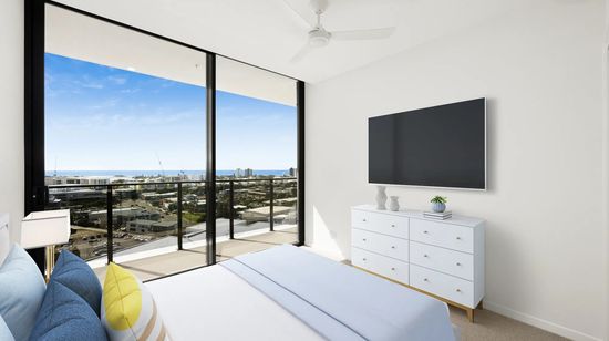 A modern bedroom in an apartment at The Corso. A neatly-made bed with several cushions faces a wall-mounted television and modern chest of drawers. The entire far wall is a full-height glass sliding door which opens out to a balcony with views of surrounding Maroochydore and the ocean in the distance.