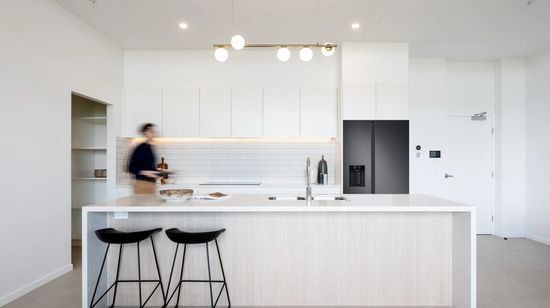 A modern kitchen in an apartment at The Corso, with a large island counter and bar standing in front of a second counter area with a stovetop and sleek refridgerator. A resident is walking through the kitchen, artistically blurred to show their motion.