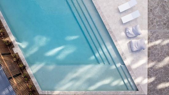 The residents’ pool at The Corso seen from above. The pool is clear blue and features full-width stairs leading down into it. Various white fabric cushions are placed on the tiled deck area along the pool’s edge.