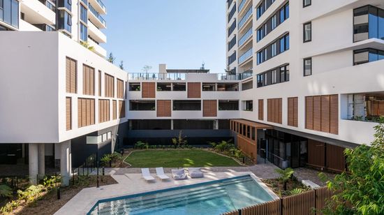 The residents’ pool and lawn area in between the buildings of The Corso. Various white fabric cushions are placed on the tiled deck area along the pool’s edge.