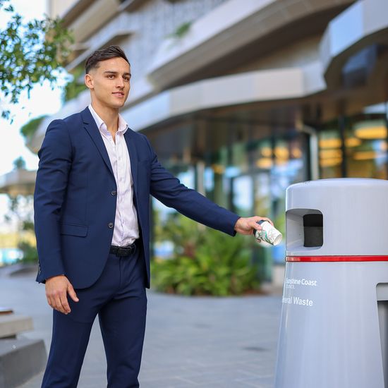 A man wearing a suit disposing of a coffee cup into a public rubbish bin labelled general waste.