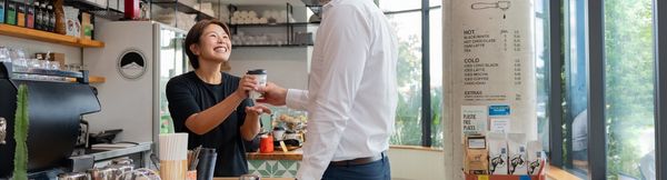 A smiling barista handing a coffee to a customer inside a bright café.